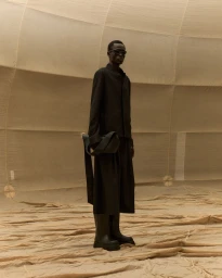 Model Daniel Ajang wearing an all-black jacket, wide trousers, sunglasses, and chunky boots against a beige fabric backdrop.