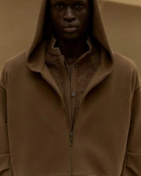 Model Daniel Ajang wearing an earthy brown hooded zip-up jacket over a collared shirt against a warm brown backdrop.