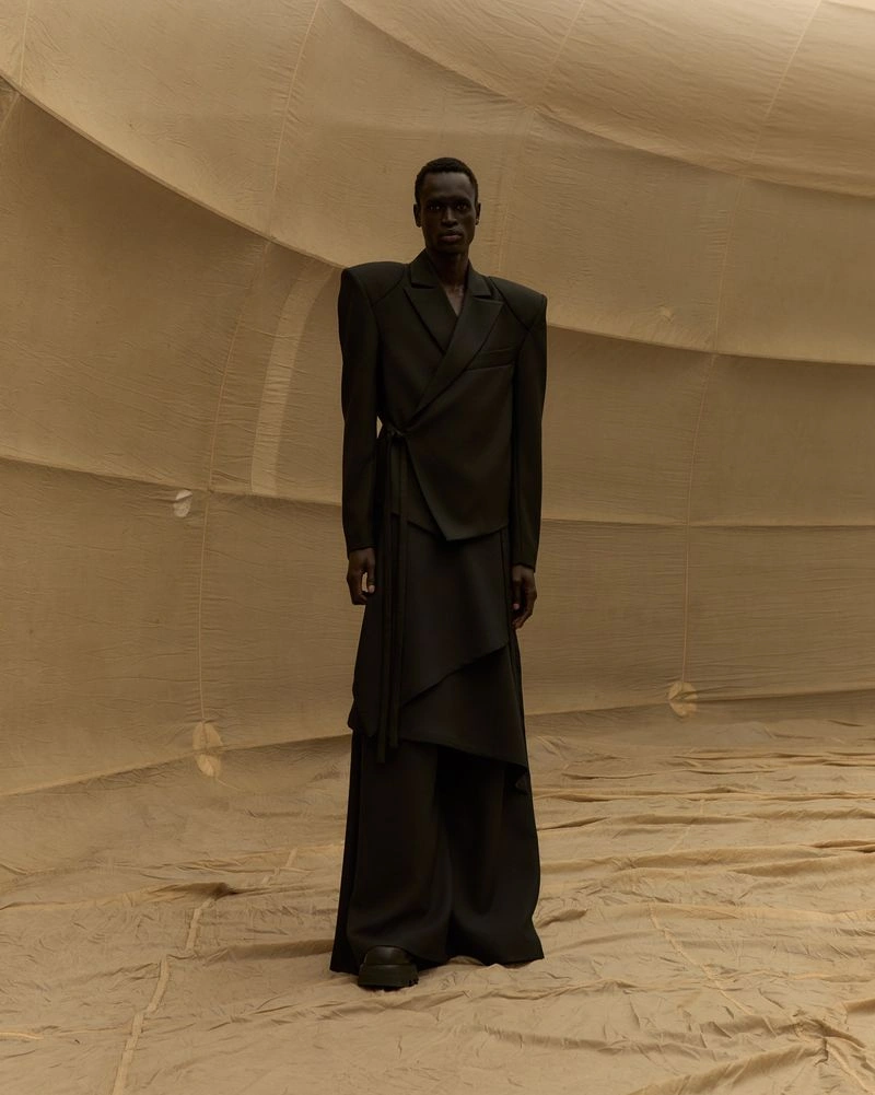 Model Daniel Ajang wears an exaggerated shoulder dark wrap suit standing inside a beige fabric enclosure for UNVAIN S/S 25.