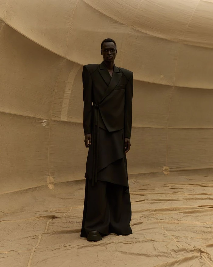 Model Daniel Ajang wears an exaggerated shoulder dark wrap suit standing inside a beige fabric enclosure for UNVAIN S/S 25.