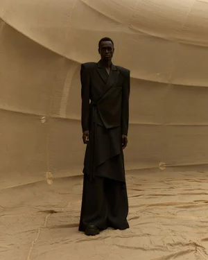 Model Daniel Ajang wears an exaggerated shoulder dark wrap suit standing inside a beige fabric enclosure for UNVAIN S/S 25.
