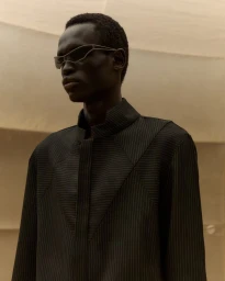 Model Daniel Ajang wearing futuristic wireframe sunglasses and a dark pinstripe coat against a warm neutral studio backdrop.