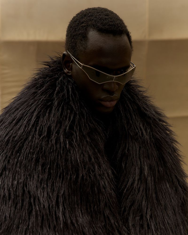 Model Daniel Ajang wearing futuristic sunglasses and an oversized black faux fur coat against a warm neutral backdrop.