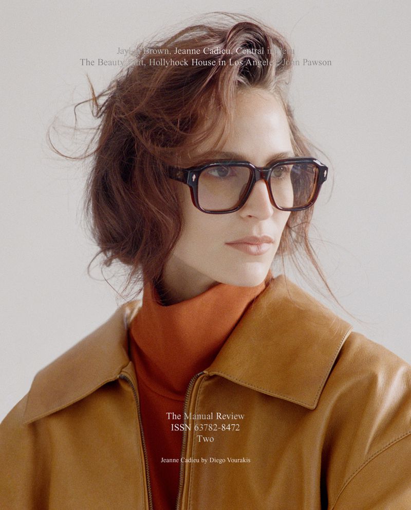 A woman with auburn hair and glasses wearing an orange turtleneck and brown leather jacket looking to her right in a studio setting.