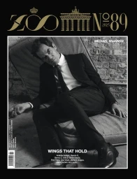 Actor Michael Shannon in a dark tailored suit and tie seated on a worn mattress against an exposed brick wall for Zoo Magazine.