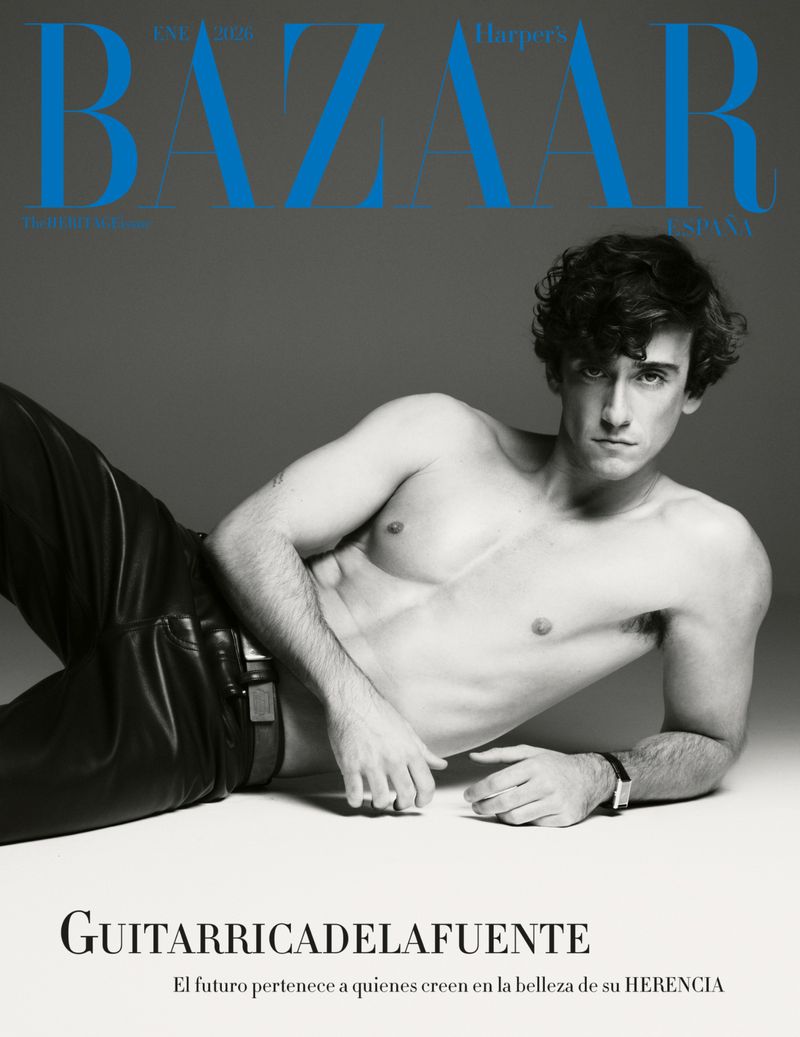 Shirtless man in black leather pants lies down for a black and white fashion editorial with 'Bazaar' magazine cover in background.