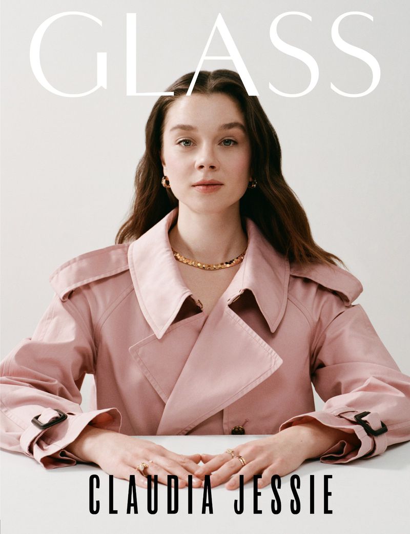 Attractive woman, Claudia Jessie, gazes forward wearing a pink trench coat and gold jewelry for Glass magazine cover; studio lighting.