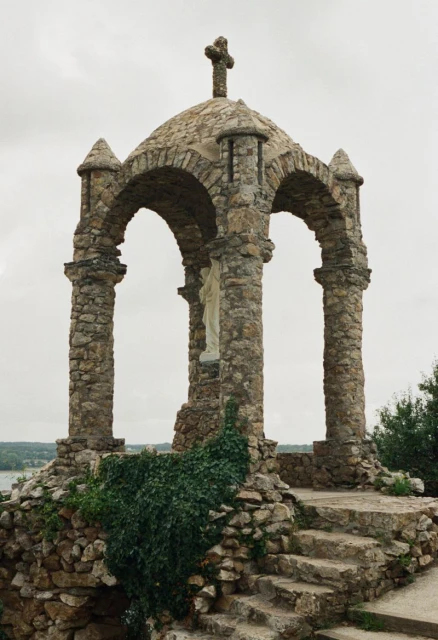 Stone gazebo with cross on top, supported by pillars, with steps leading up. Greenery surrounds the base, set against a cloudy sky.