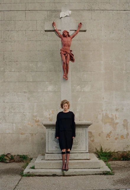 Woman in black dress stands near a stone cross with a reddish statue of Jesus in an outdoor, grey stone setting, creating a solemn mood.