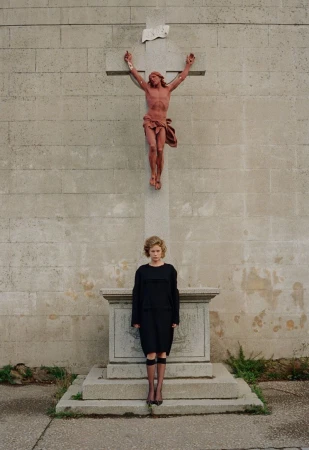 Woman in black dress stands near a stone cross with a reddish statue of Jesus in an outdoor, grey stone setting, creating a solemn mood.