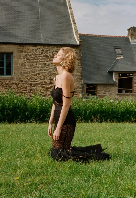 Woman in black dress kneeling in green field with stone building backdrop, bathed in sunlight. Fashion editorial in outdoor setting.