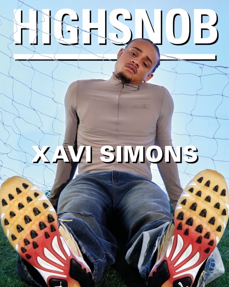 Xavi Simons poses outdoors wearing jeans and a beige long-sleeved top, with sneakers in the foreground, against a net backdrop.