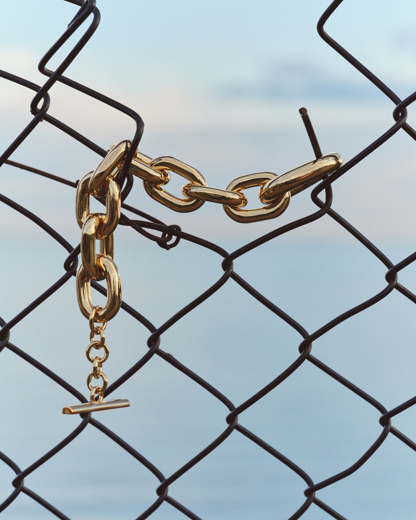 Gold chain necklace draped over a wire fence against a blurred sky background. Jewellery fashion still life, accessories shot.