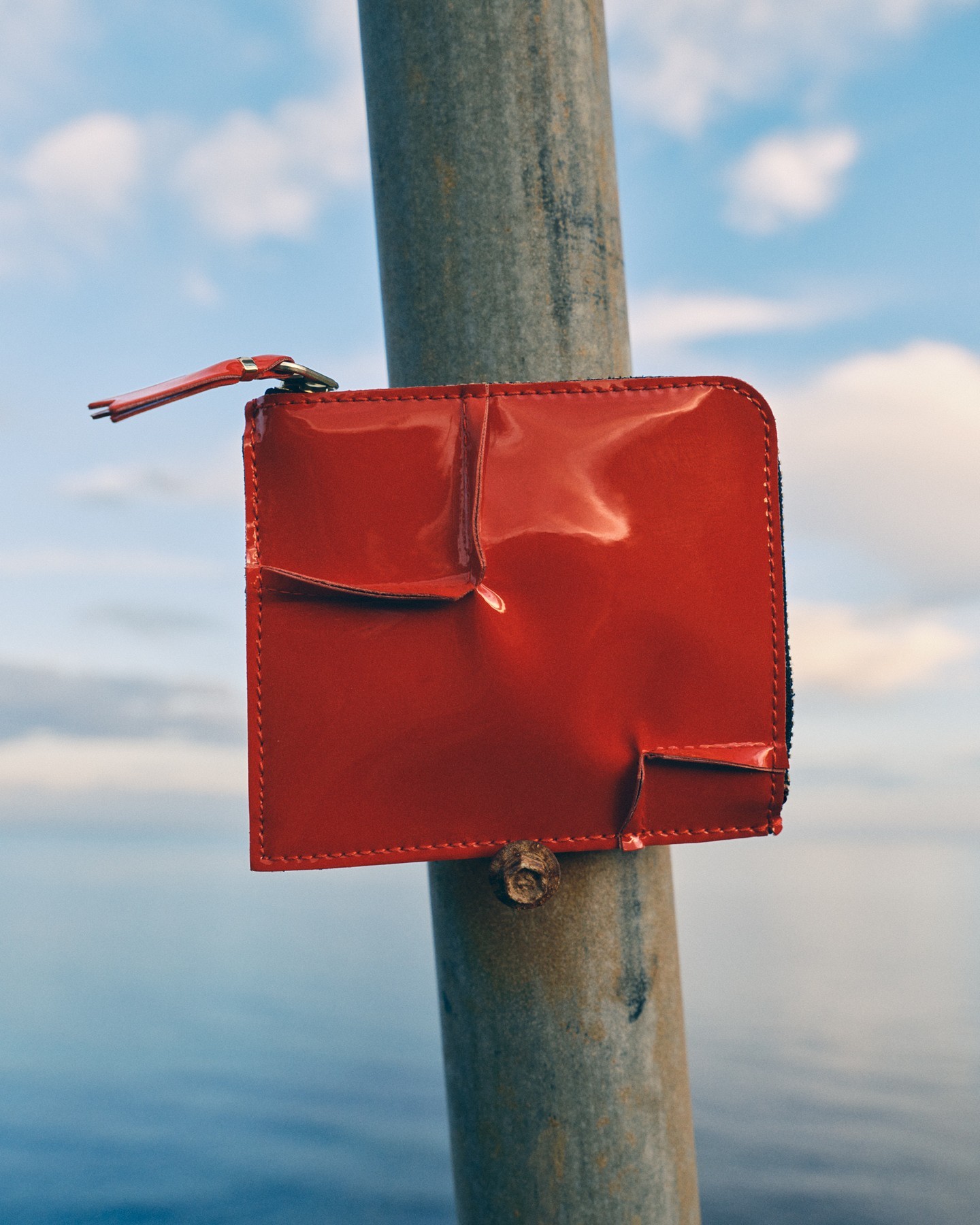 Close-up of a red, shiny wallet attached to a weathered metal pole with a blurred background of water and sky providing context.
