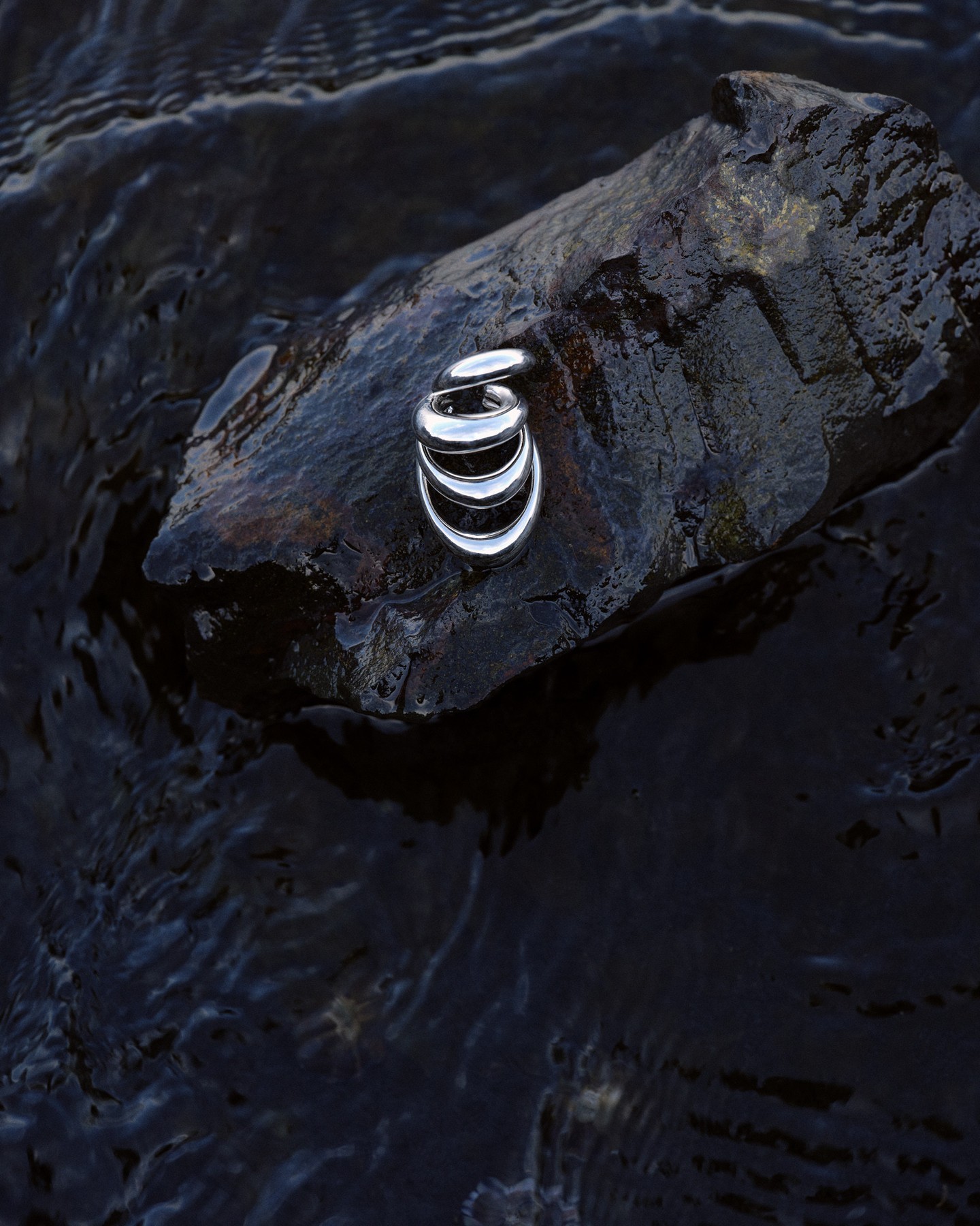 Polished silver sculptural jewellery piece resting on a wet dark rock surrounded by swirling water.