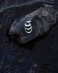 Polished silver sculptural jewellery piece resting on a wet dark rock surrounded by swirling water.