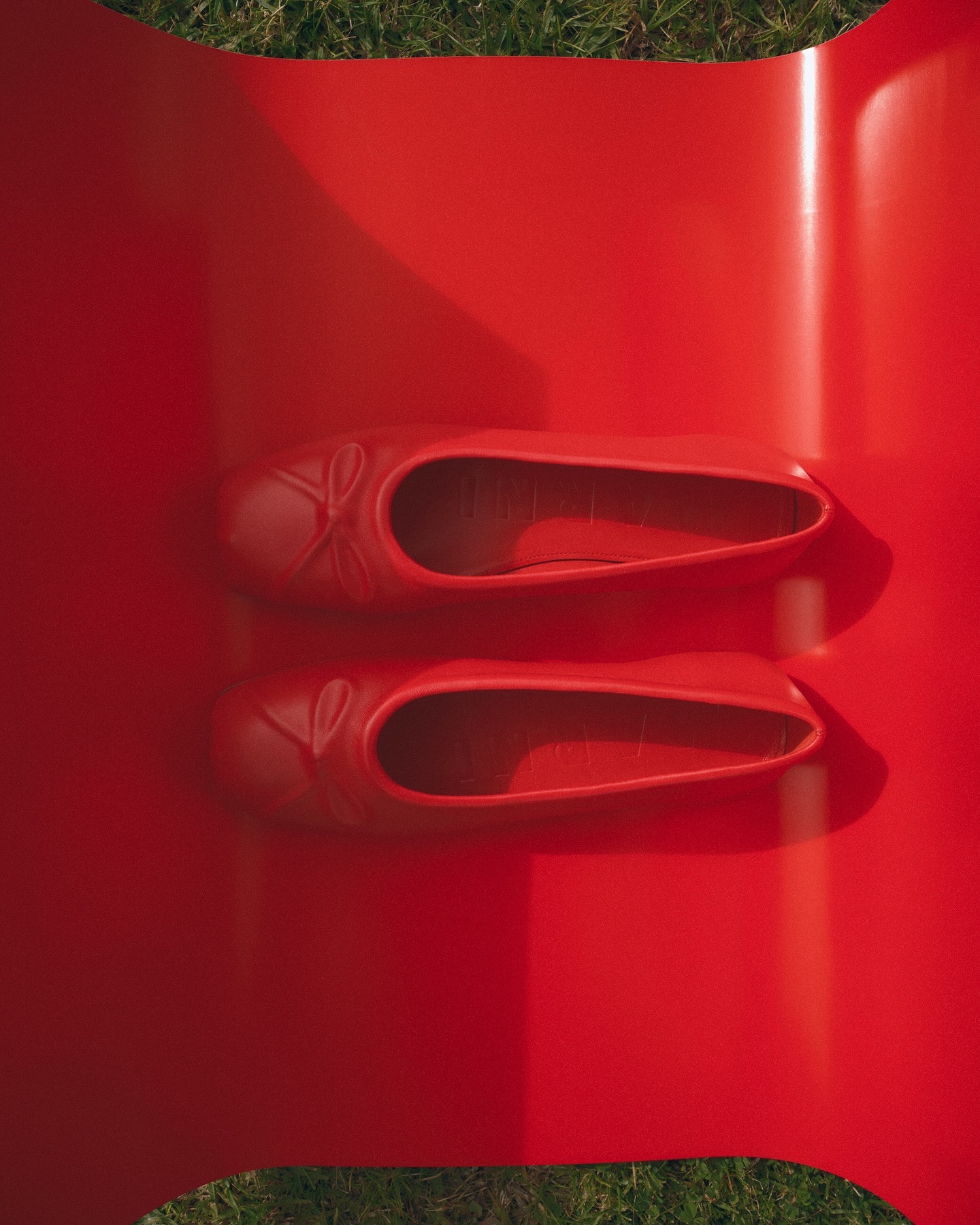 Overhead shot of two red ballet flat shoes on a red platform, set against green grass. Focus on color, texture, and material.