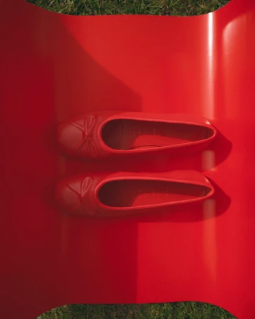 Overhead shot of two red ballet flat shoes on a red platform, set against green grass. Focus on color, texture, and material.