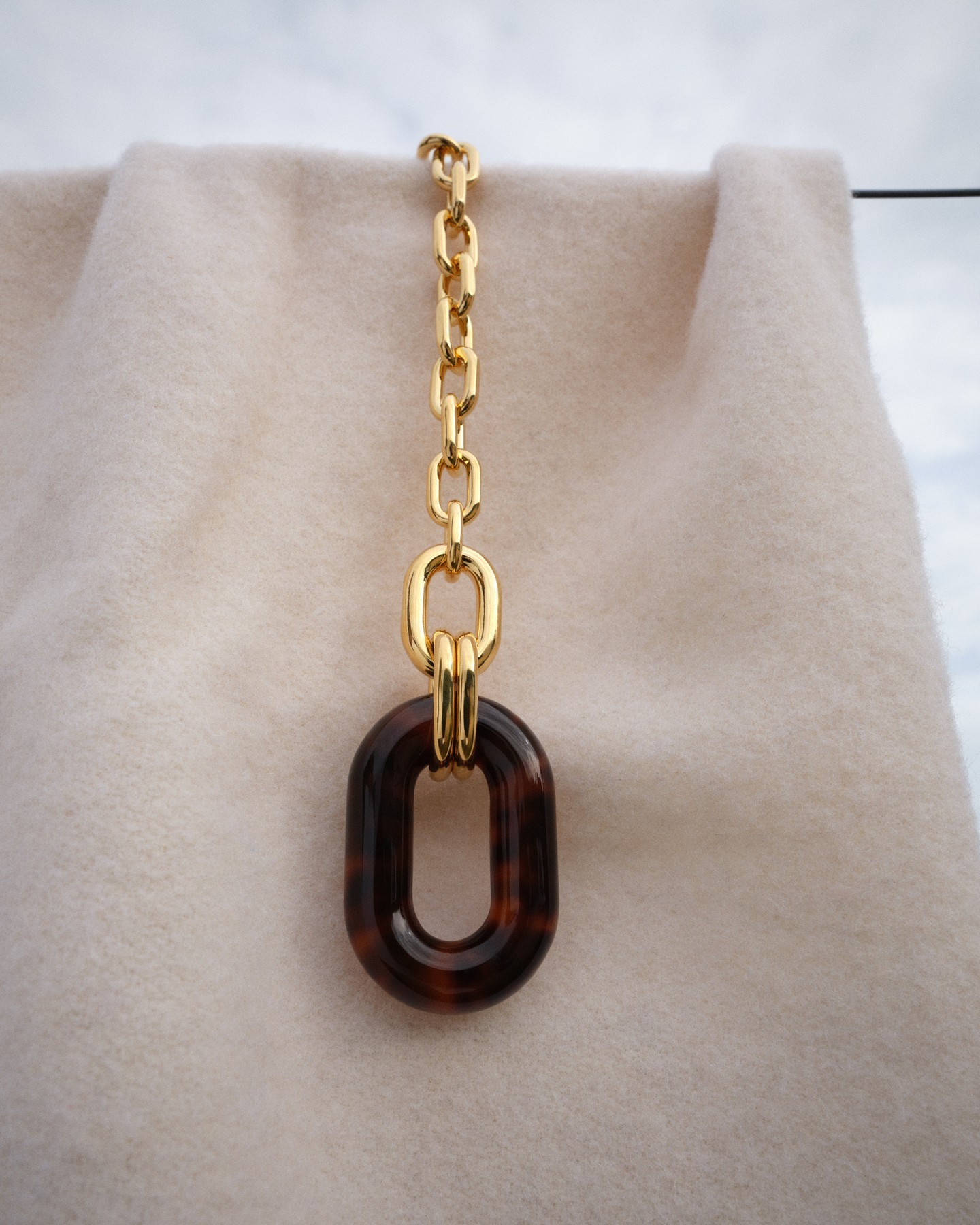 Gold chain necklace featuring a brown tortoiseshell resin link, set against a light cream-colored fabric backdrop and soft natural light.