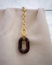 Gold chain necklace with tortoiseshell pendant draped over a cream-colored textured textile against an overcast sky.