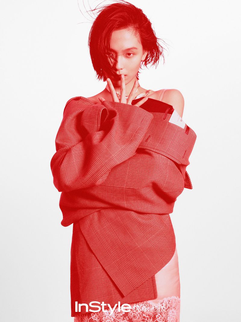 Fashion editorial shot of a young adult woman with short hair wearing a red blazer and lace skirt against a light background.