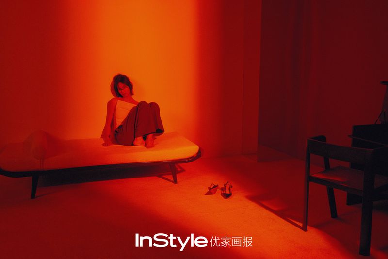 A woman with short hair is sitting on a sofa in a room with a red color scheme. Two high heels sit on the floor near the sofa.