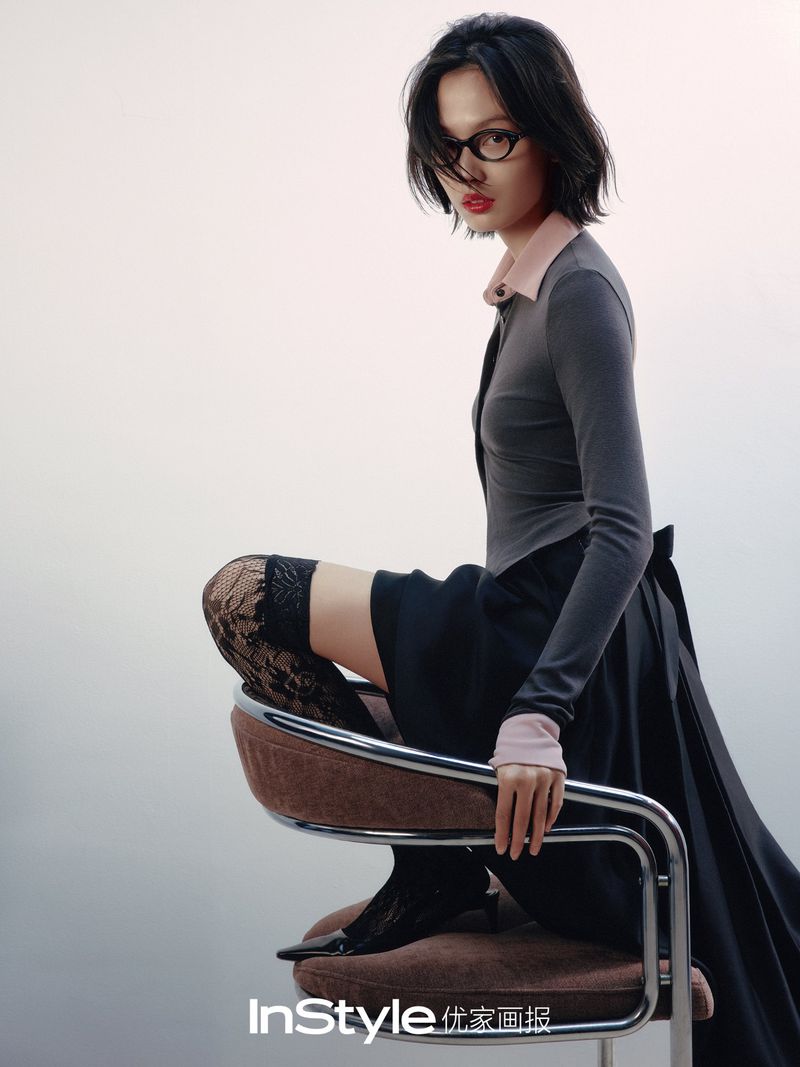 A stylish woman with short black hair and glasses poses in gray top, black skirt, lace stockings on a brown chair in a light studio.