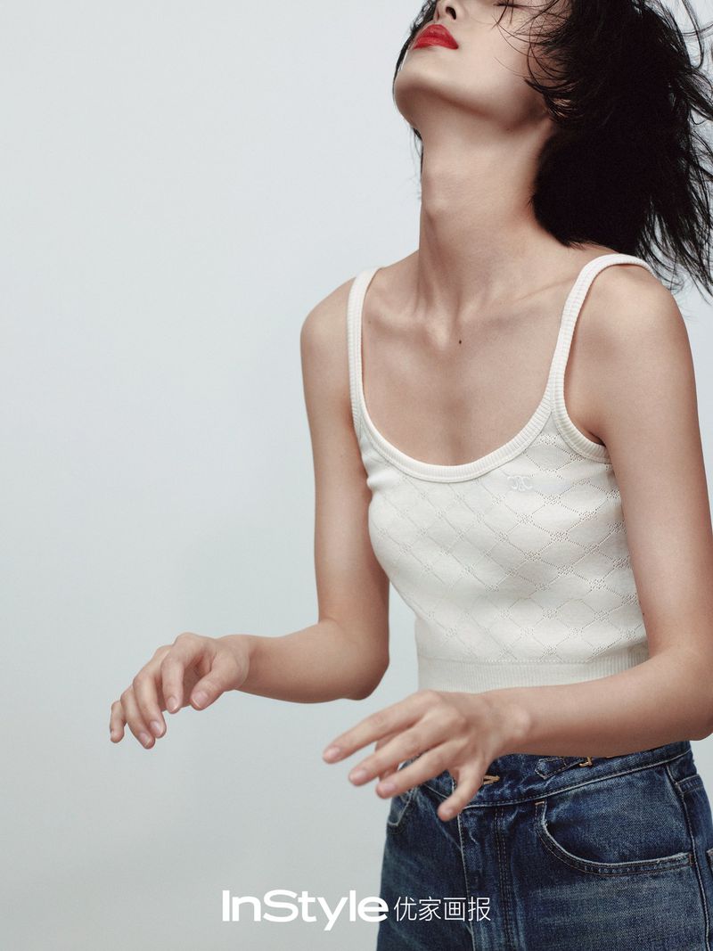 A woman in a white tank top and blue jeans tilts her head back slightly against a white background in a fashion editorial shot.