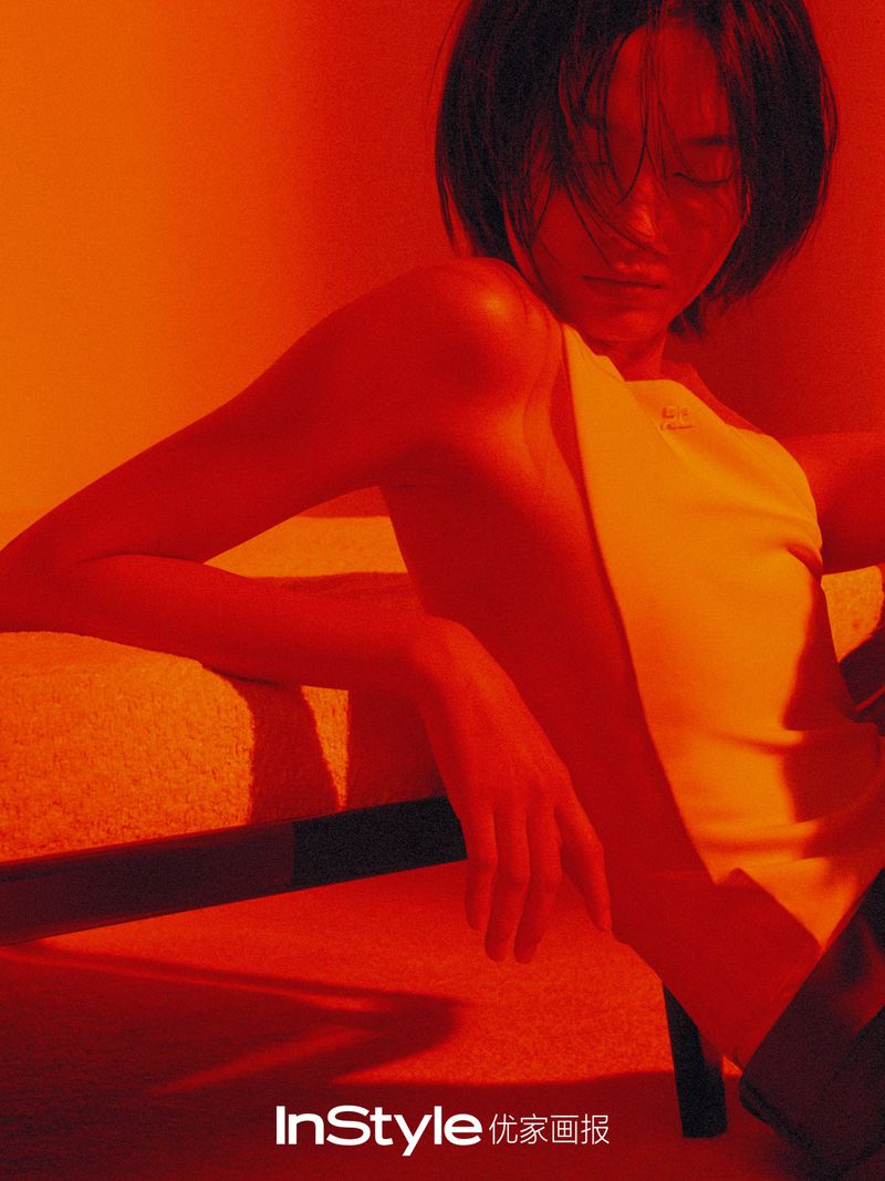 Portrait of an adult woman with short dark hair in a white top, leaning on an orange surface under orange-red lighting. 'InStyle' logo visible.