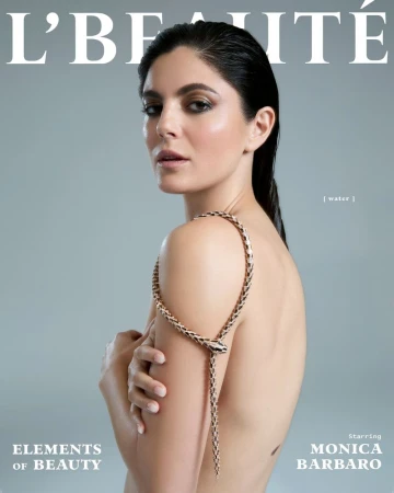 Close-up magazine cover featuring Monica Barbaro, a brunette woman with bare shoulders, wearing a snake-shaped necklace against a gray background.