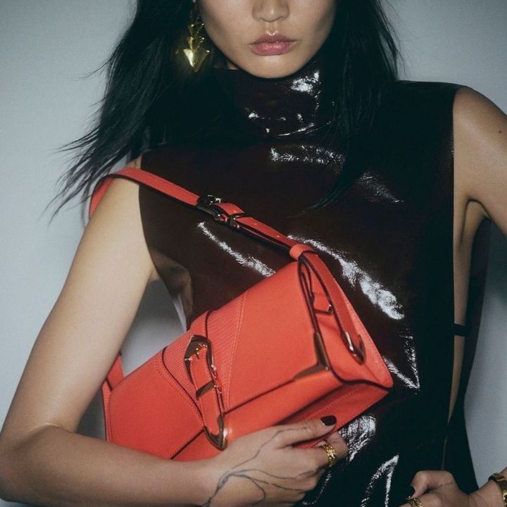 Model wearing a shiny dark brown top and holding a bright orange structured handbag with a buckle.