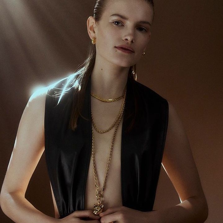 Woman poses in a sleeveless black top, wearing multiple gold necklaces in a studio with soft lighting, highlighting her jewelry.