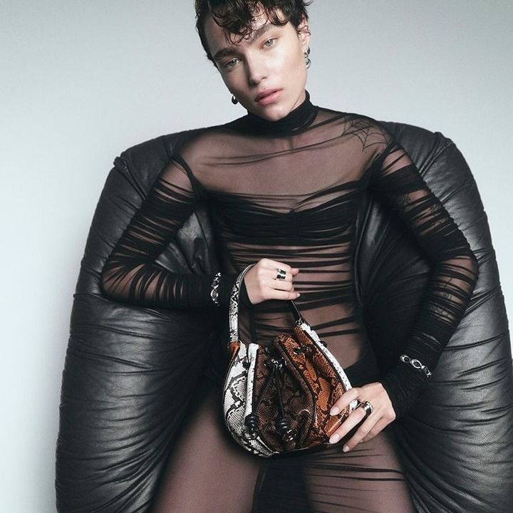 A slender person with short dark hair poses in a black sheer top and skirt, holding a snakeskin-patterned purse while sitting in a black chair.