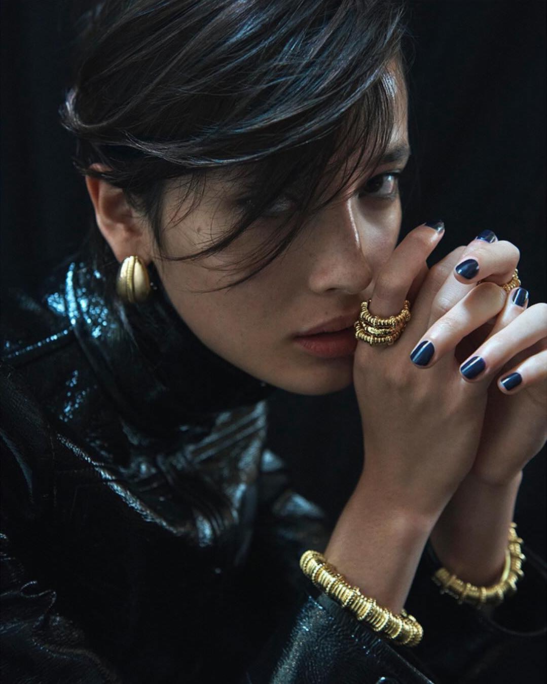 A woman with dark hair poses in a close-up fashion shot, wearing gold jewelry: earrings, rings, and a bracelet, against a black background.
