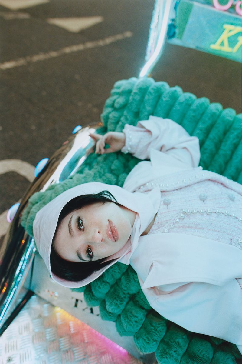 Young woman in a hooded pink jacket reclining on a green faux-fur surface, creating an eye-level, medium indoor portrait.