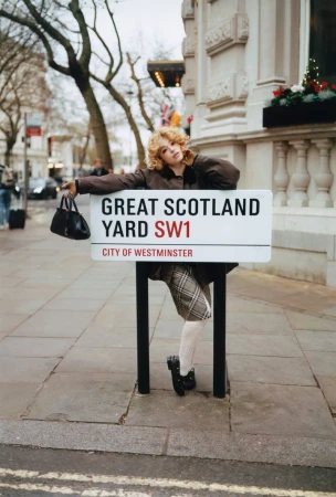 Woman poses with Great Scotland Yard street sign in London. She wears a blazer, skirt, tights, and heels, holding a black purse.
