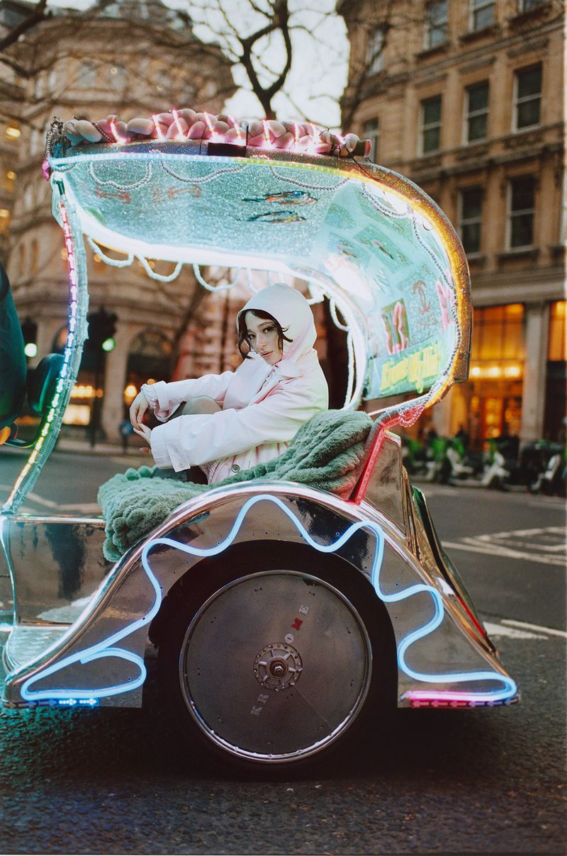 Young woman in light pink coat with hood riding in a decorated auto rickshaw on an urban street with buildings in the background.