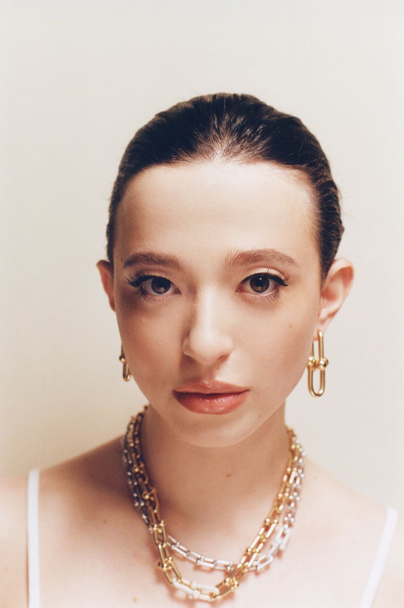 Portrait of Mikey Madison wearing layered link necklaces and gold hoop earrings against a pale background.