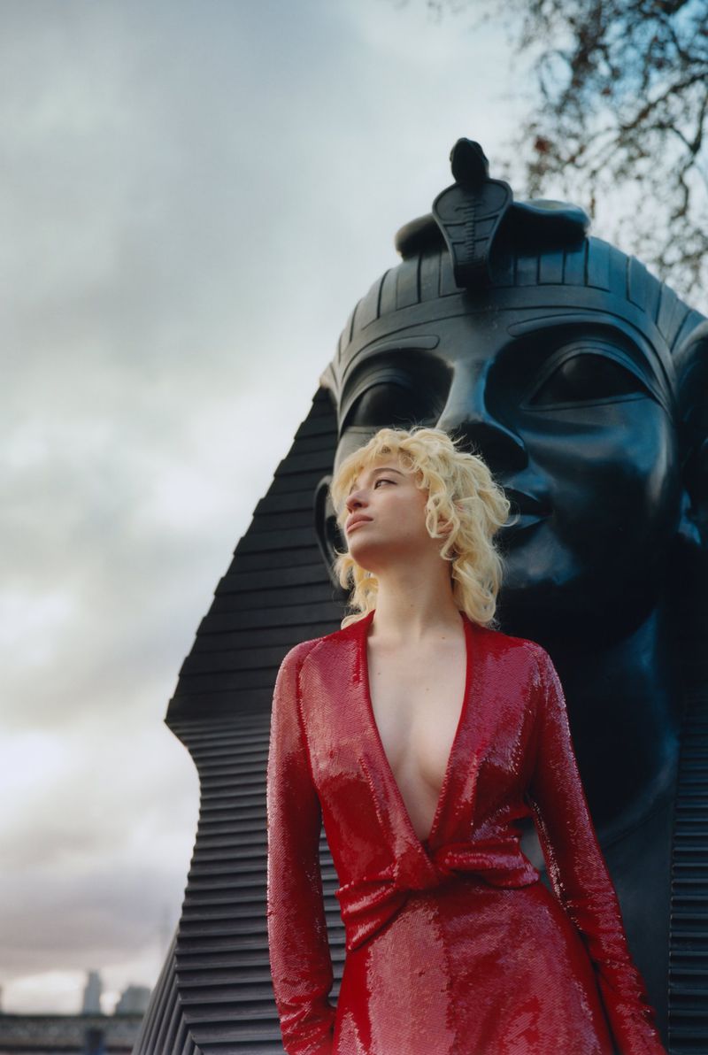 Blonde woman in red dress stands before a colossal black sphinx sculpture under a cloudy sky, blending fashion with ancient art.