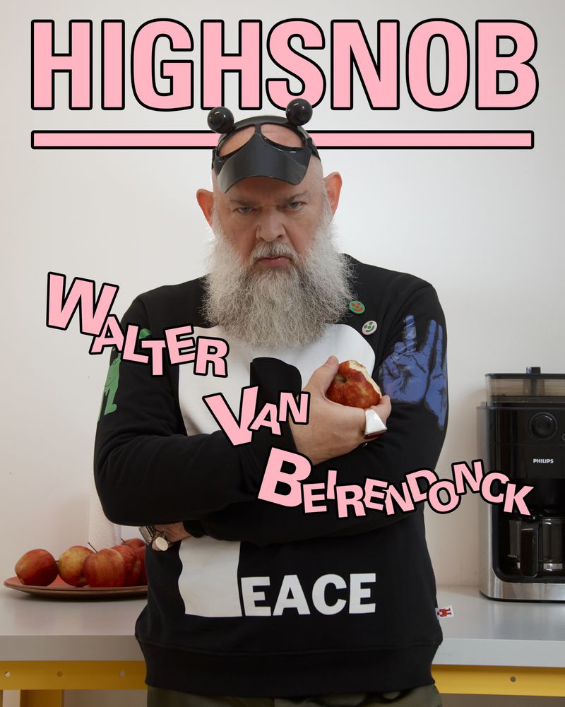 Man with a long beard poses in a black sweater with 'PEACE' on it, holding an apple in a modern kitchen with white walls.