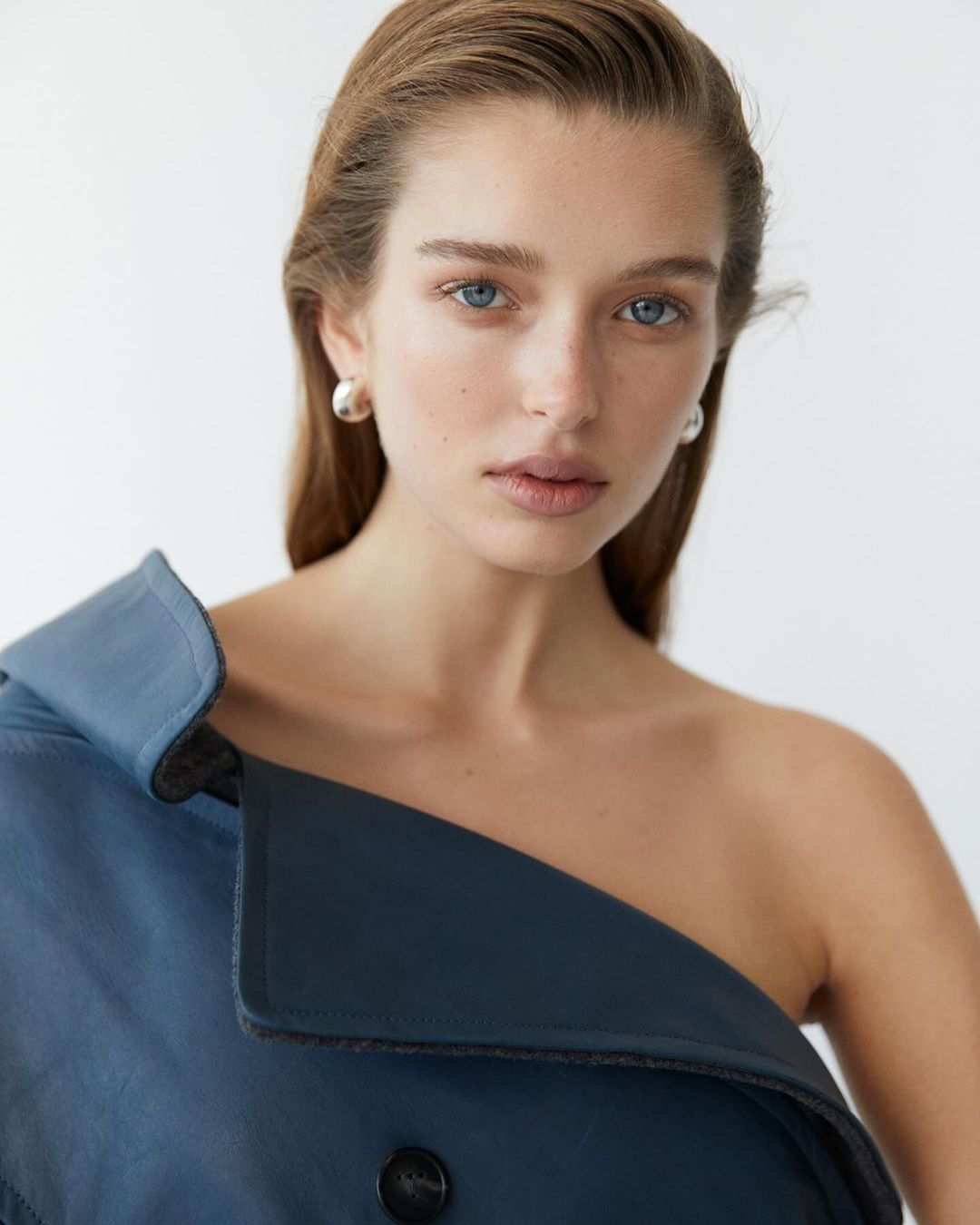 Close-up portrait of a young fair-skinned woman with blue eyes, wearing an off-the-shoulder blue coat against a white background.