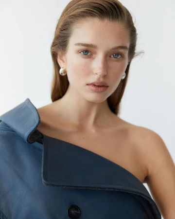 Close-up portrait of a young fair-skinned woman with blue eyes, wearing an off-the-shoulder blue coat against a white background.