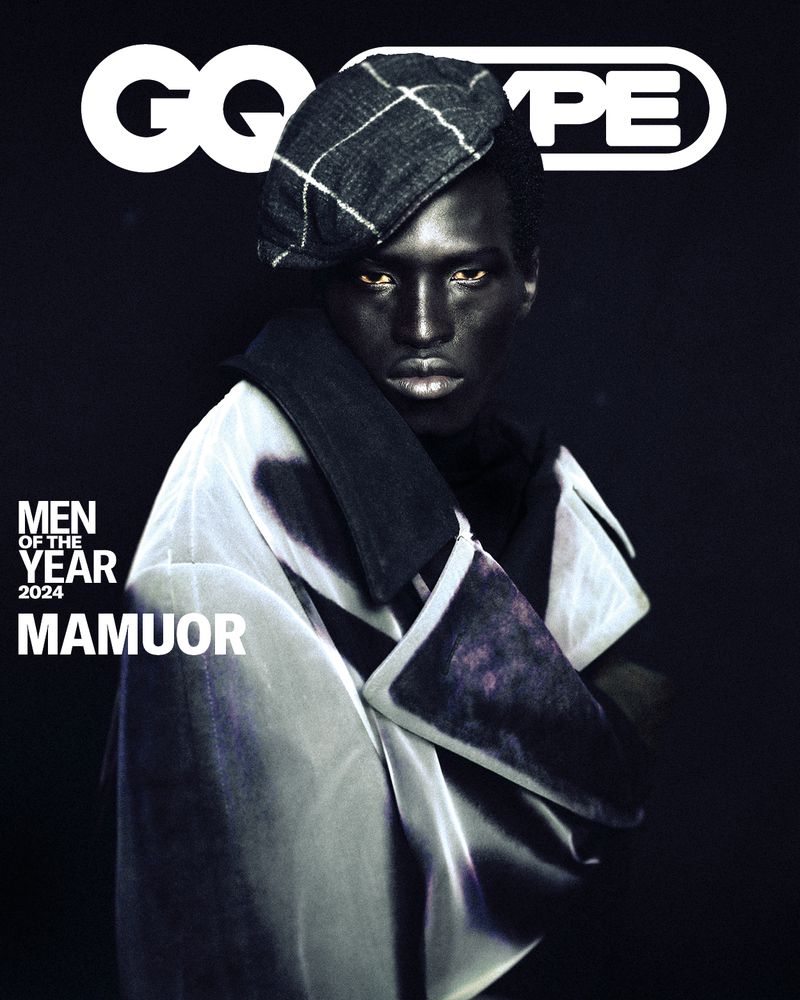 Portrait of Black man, Mamour, wearing a beret and draped coat, GQ cover with black backdrop. Fashion editorial portrait.