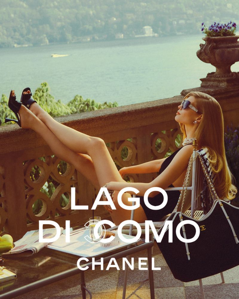 A blonde woman reclines on a chair in black swimwear and sunglasses, with a handbag beside her, with Lake Como and mountains.