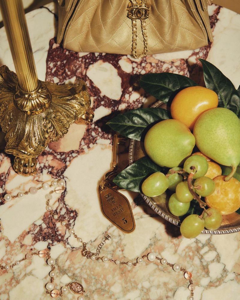 Still life of an expensive quilted gold purse, fruit, a hotel keychain, and pearl necklace on a marbled surface in warm lighting.