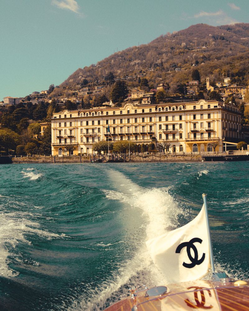 A scenic view of a grand building on a lakeshore, captured from a boat with a branded flag, set against a lush mountain backdrop.