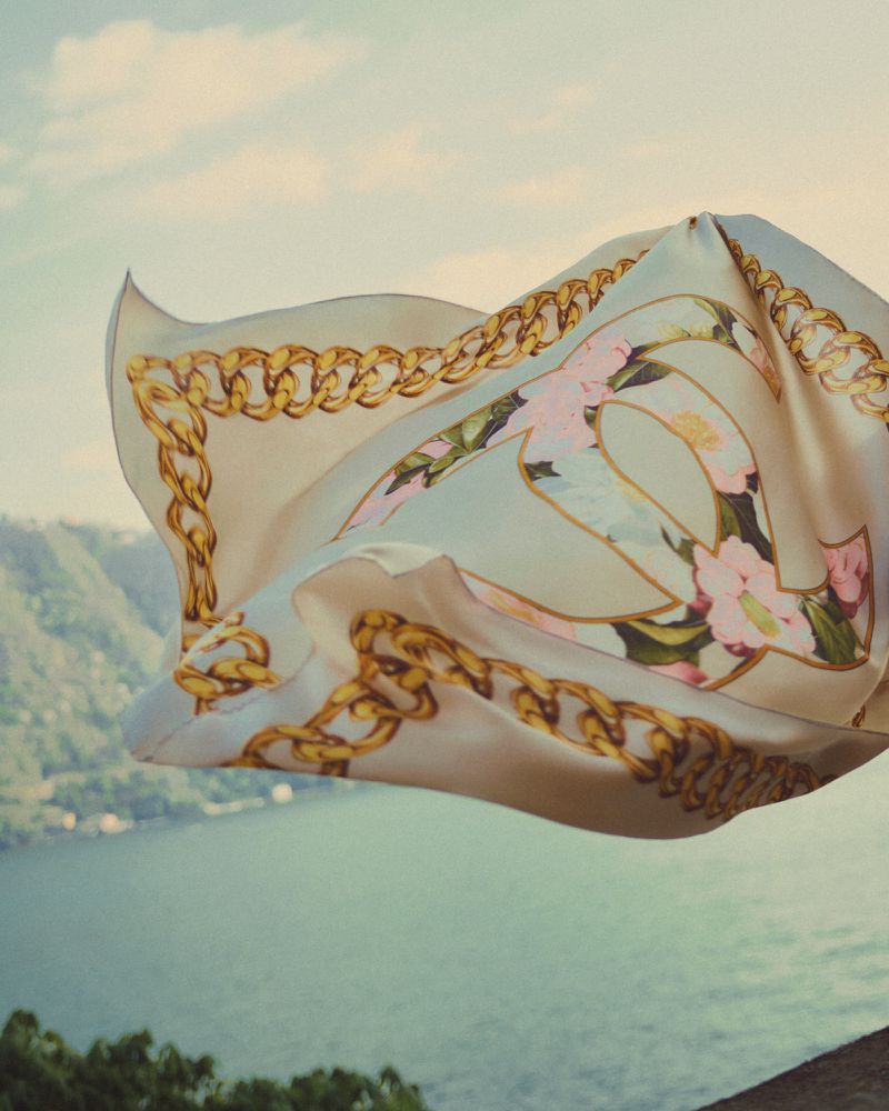 Silk scarf with gold chain pattern and floral motifs fluttering in the wind above a large body of water and distant hills.