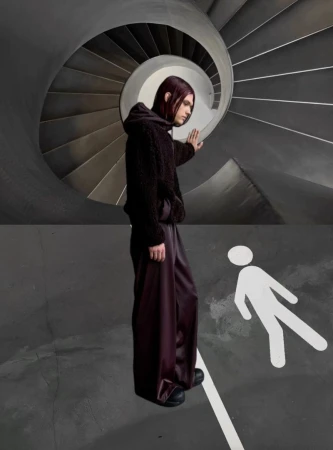 A person stands in front of a pedestrian crossing symbol and a spiraling staircase in an abstract studio-lit fashion shot.