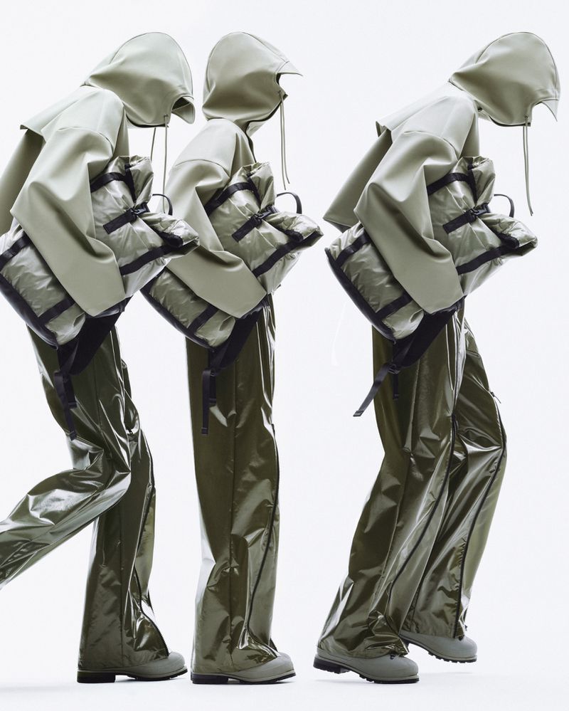 Three figures in olive green hooded raincoats and trousers, each carrying a matching bag, against a bright white backdrop.