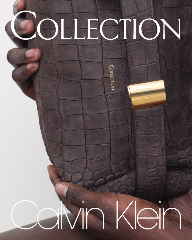 Close up of a brown leather Calvin Klein purse with gold hardware held by a person in front of a light gray background.